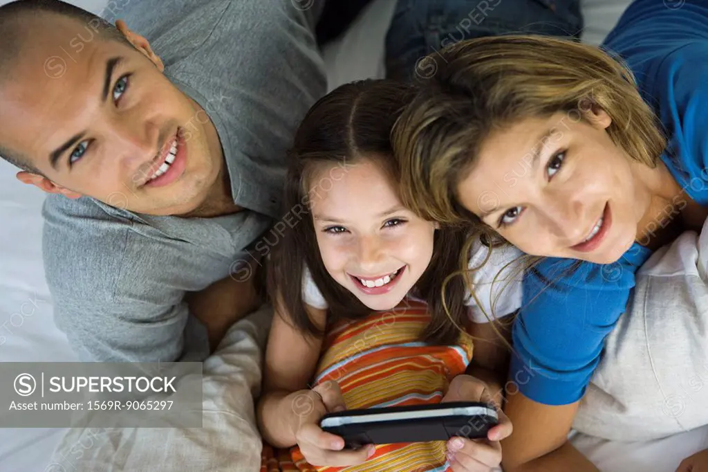 Girl lying between parents, playing handheld video game, all smiling up at camera