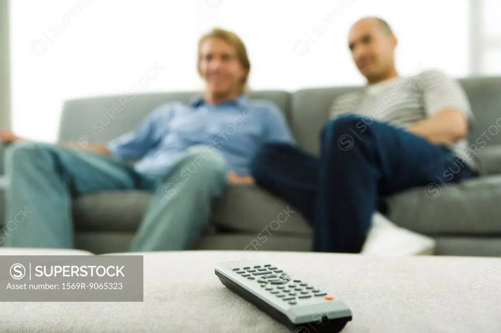 Two men sitting on sofa, focus on remote control in foreground