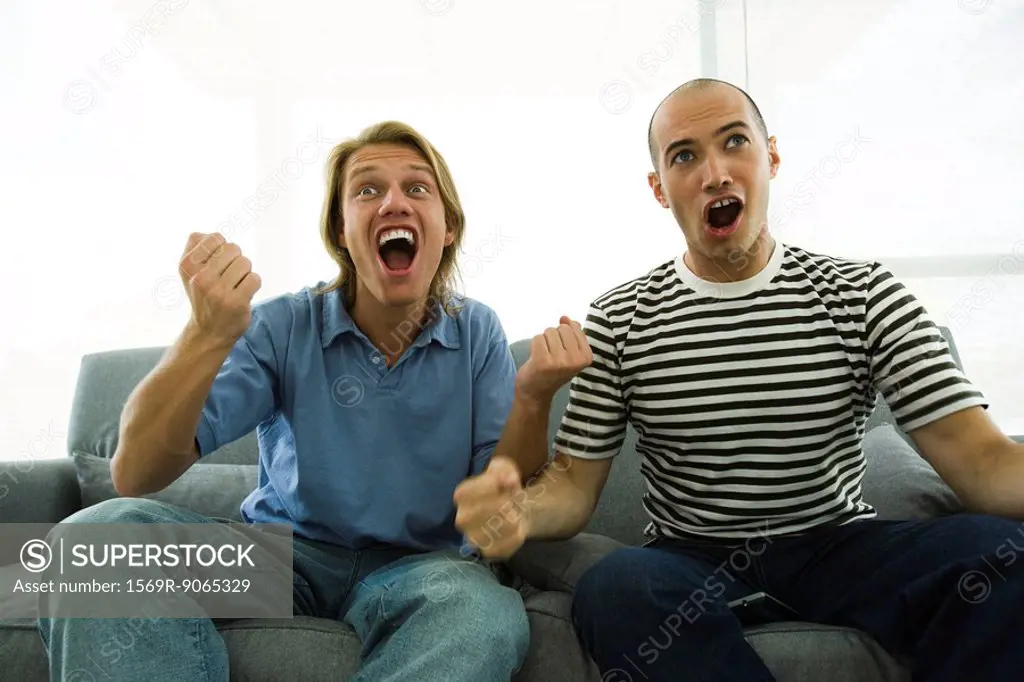 Two men sitting on sofa watching TV, cheering with clenched fists
