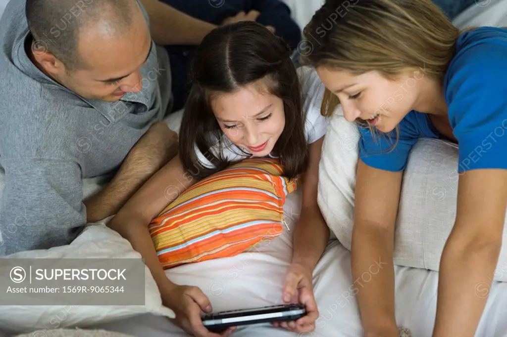 Girl lying with parents, playing handheld video game