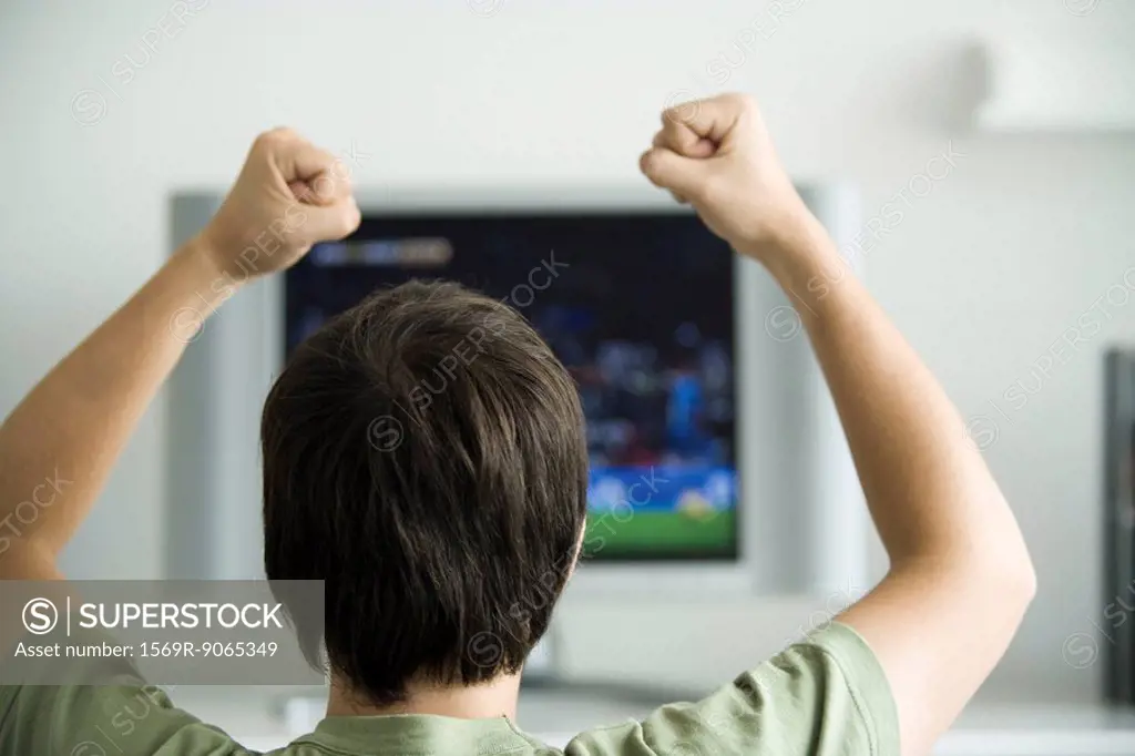 Male watching television, fists raised in air, rear view