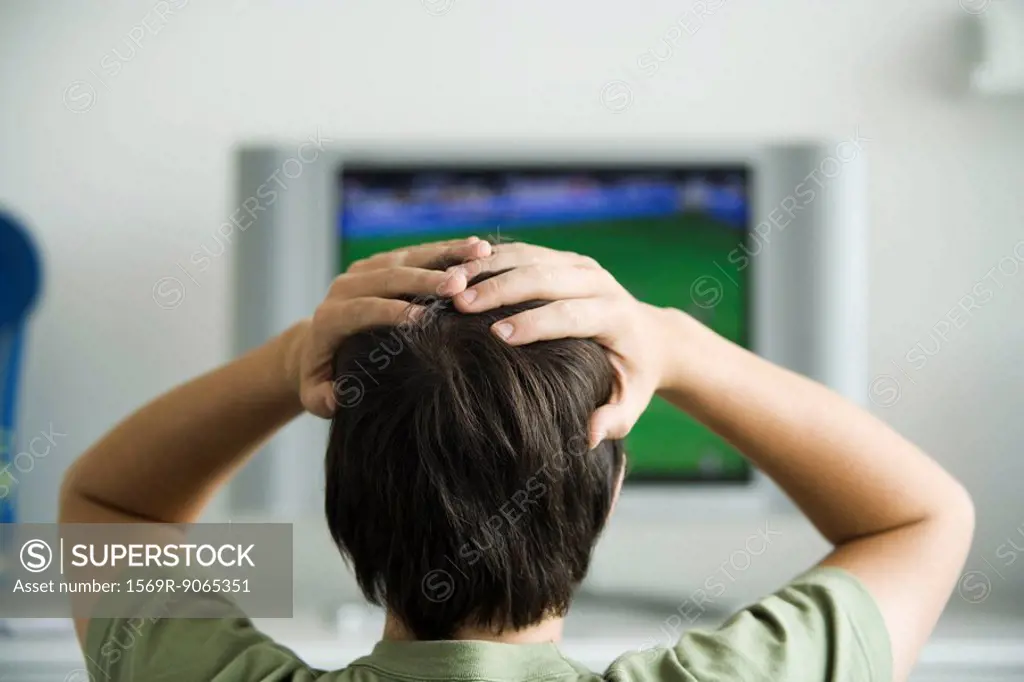 Male watching television, both hands on head, rear view