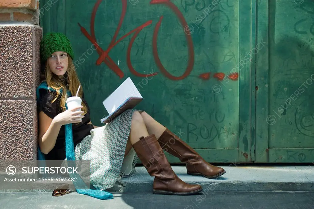 Young woman sitting on ground with book and drink, looking away