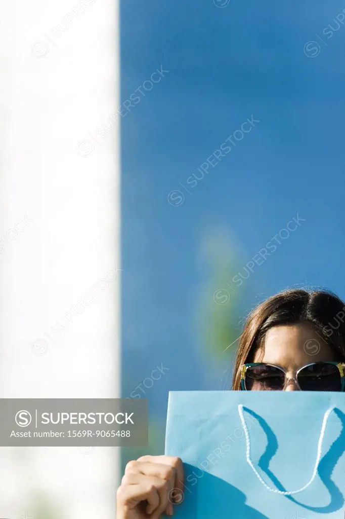 Young woman wearing sunglasses, holding shopping bag in front of face
