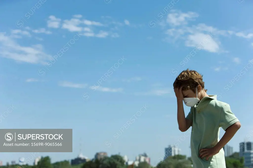 Boy standing outdoors wearing pollution mask, holding head, city in background