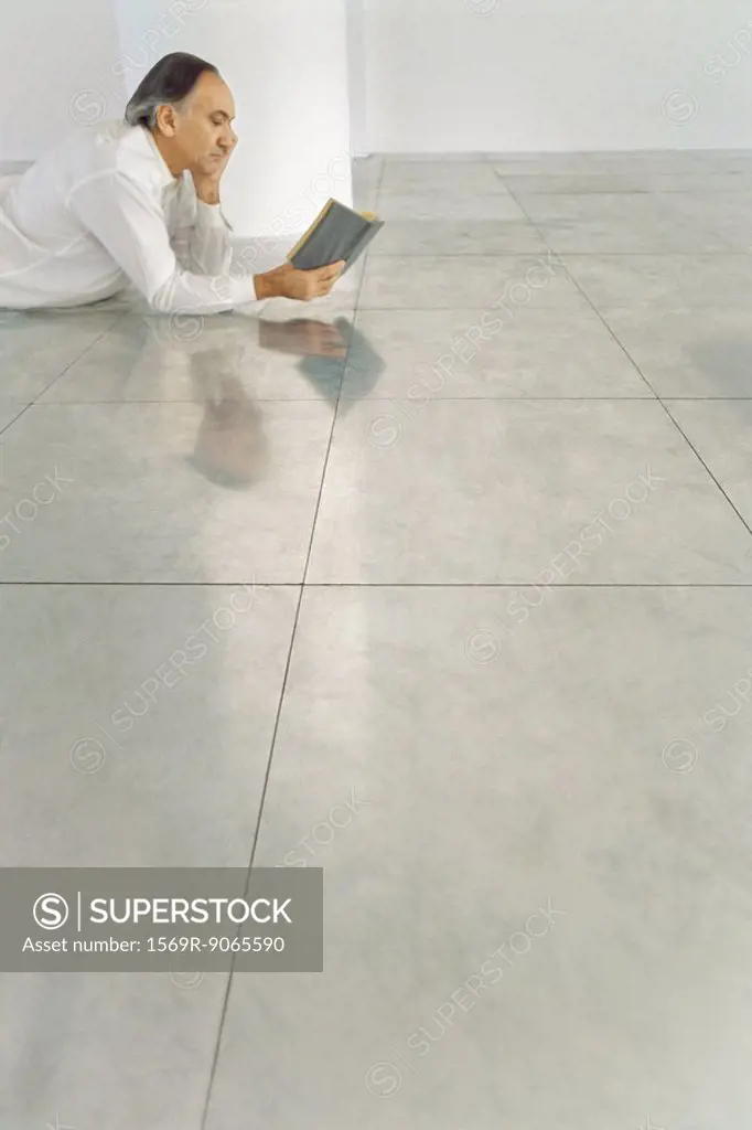 Senior man lying on stomach on tiled floor reading book