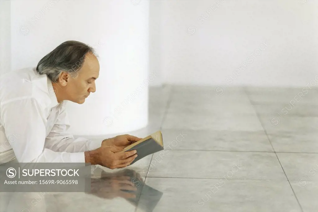 Senior man lying on tiled floor reading book