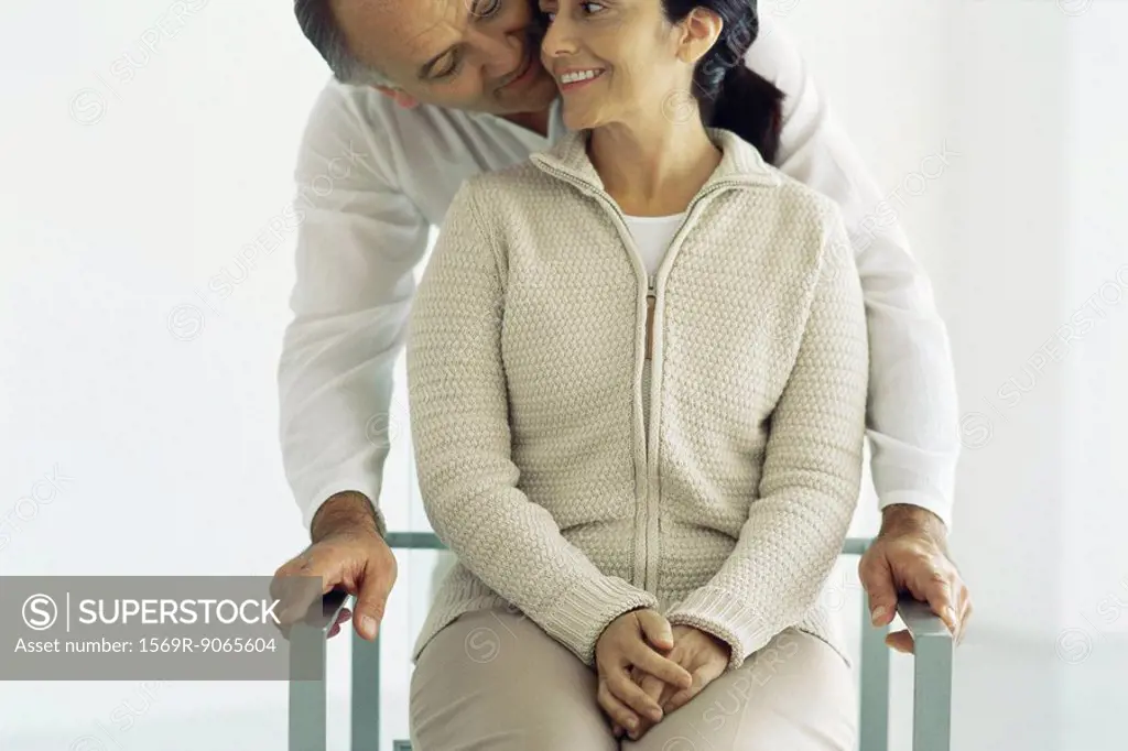 Woman sitting in chair, husband standing behind leaning down whispering in her ear, front view