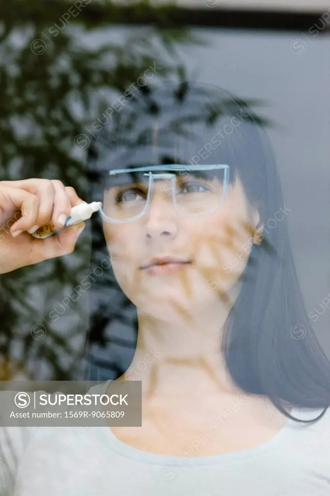 Young woman drawing a pair of glasses on a window pane, looking through window
