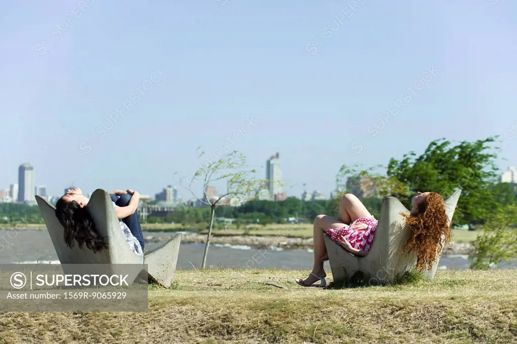 Two young women relaxing in chairs, pond and city skyline in background