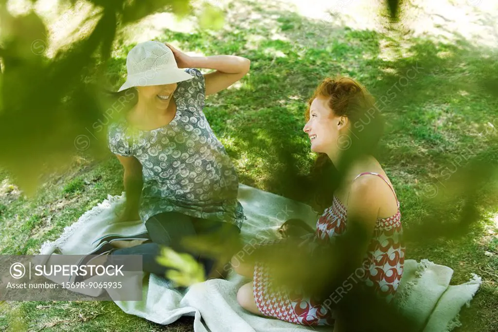 Two young women sitting on blanket, talking, viewed through foliage