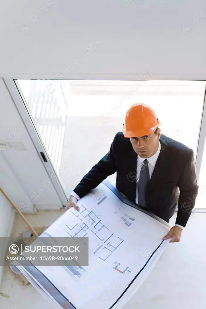 Man in suit wearing hard hat holding blueprints looking up at camera