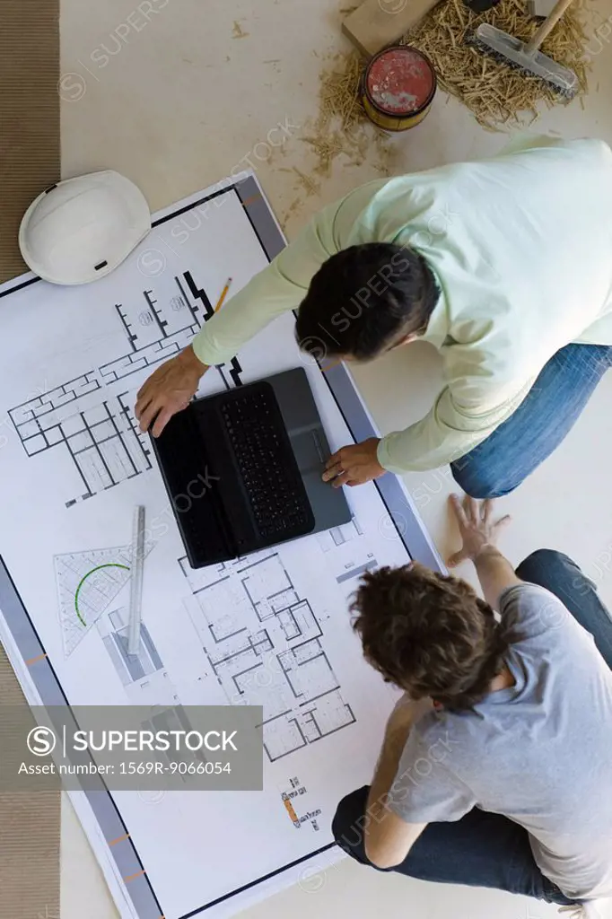 Two men crouching looking over blueprints, using laptop set on floor