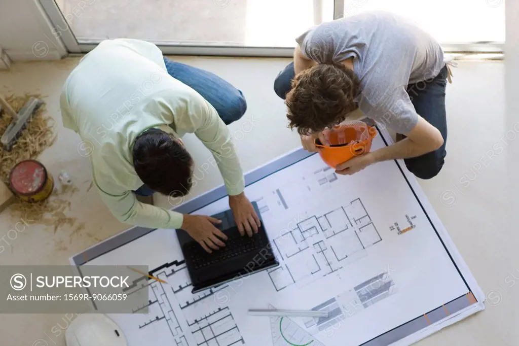 Two men crouching using laptop, looking at blueprint spread out on floor