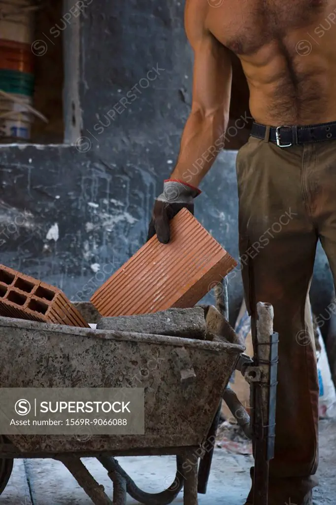 Barechested construction worker placing brick in wheelbarrow