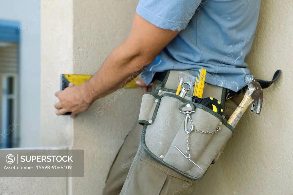 Construction worker wearing tool belt heavy with tools measuring wall