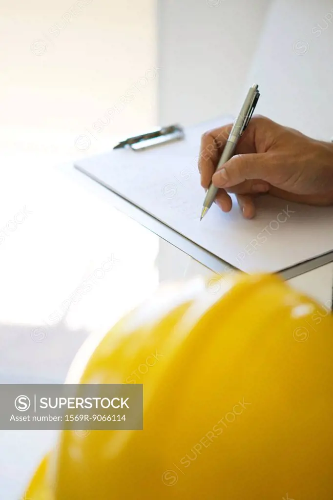 Hand writing notes on clipboard pad, hard hat in foreground, cropped view