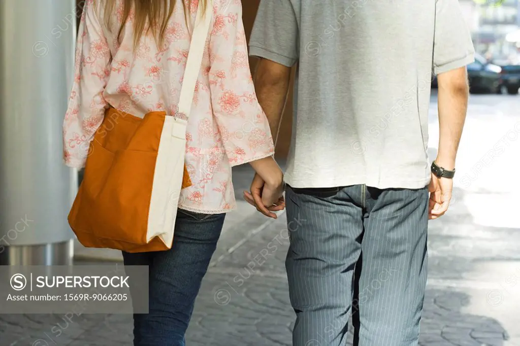Young couple walking hand in hand on sidewalk, rear view, cropped