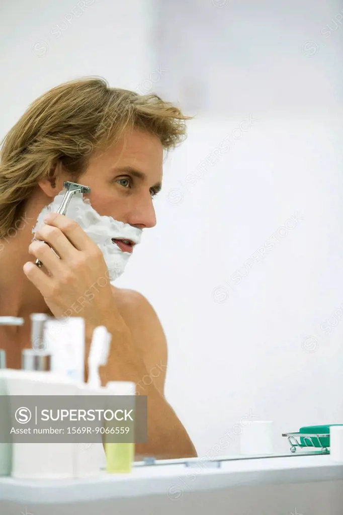 Man shaving with razor, looking at self in bathroom mirror