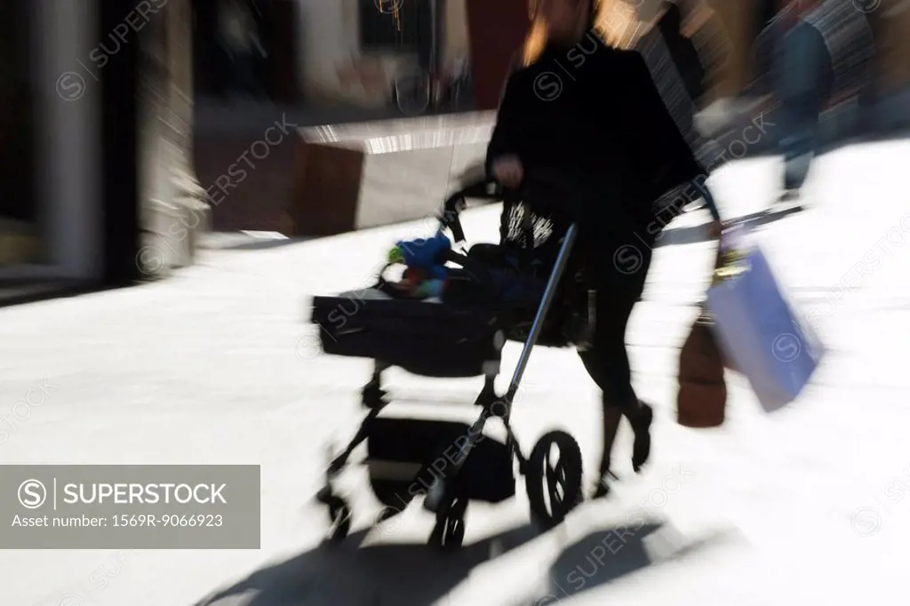 Pedestrian pushing baby carriage on sidewalk, carrying shopping bags, blurred