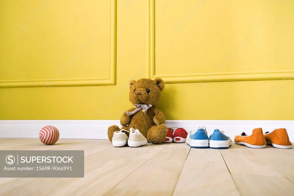 Teddy bear sitting on floor with several pairs of shoes, ball nearby