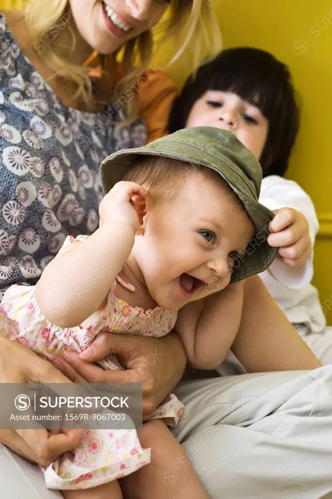 Infant girl sitting with mother and brother, wearing hat and laughing