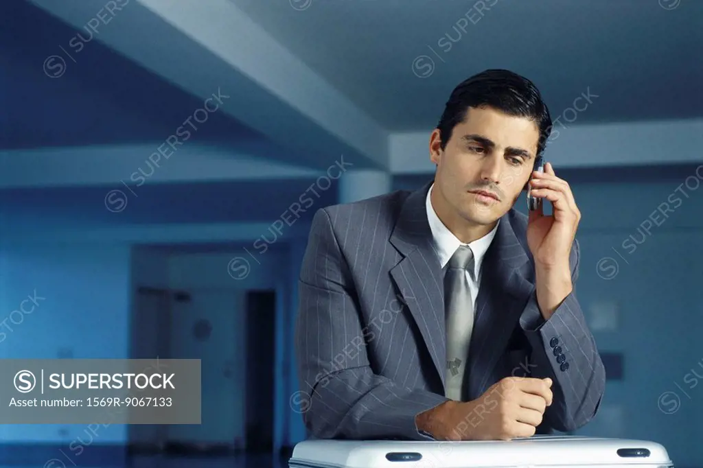Businessman sitting with briefcase on lap in empty office using cell phone