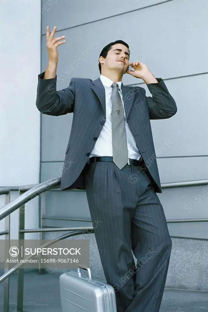 Businessman leaning against railing and using cell phone, making frustrated gesture with hand