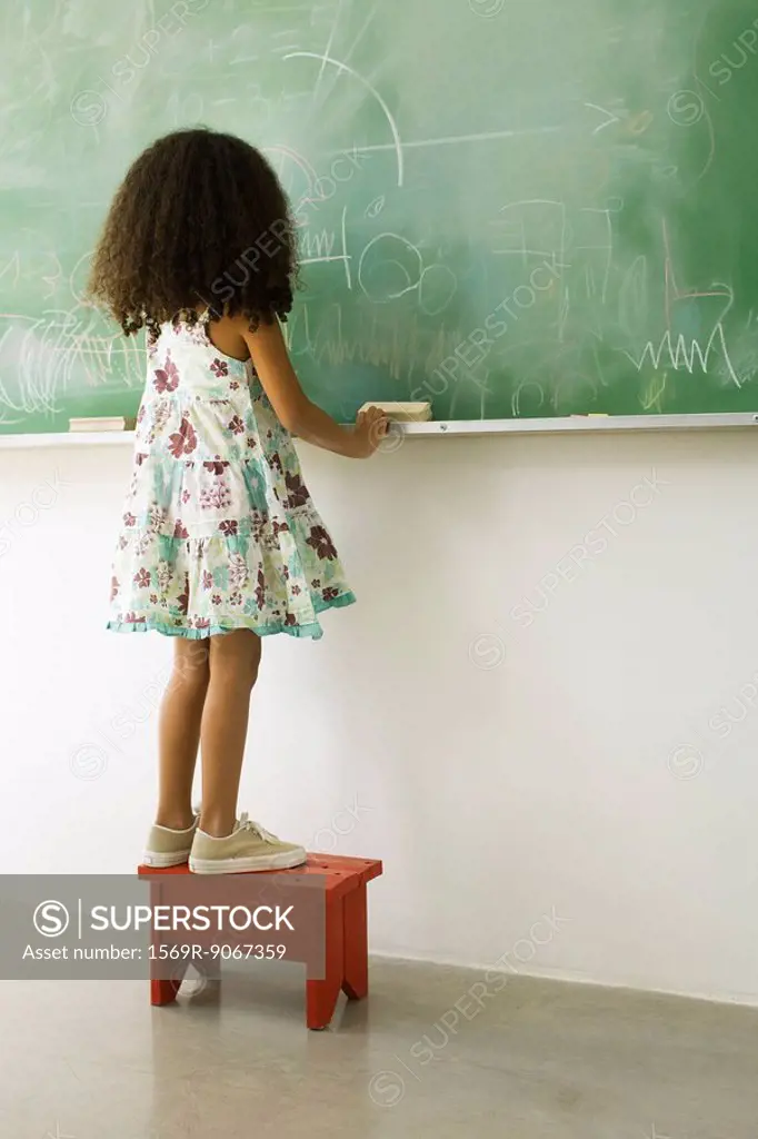 Little girl standing on stool in front of blackboard, rear view