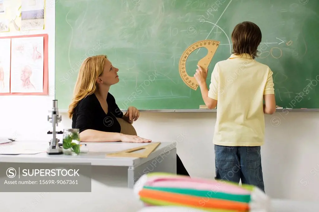 Boy standing in front of classroom blackboard, using protractor