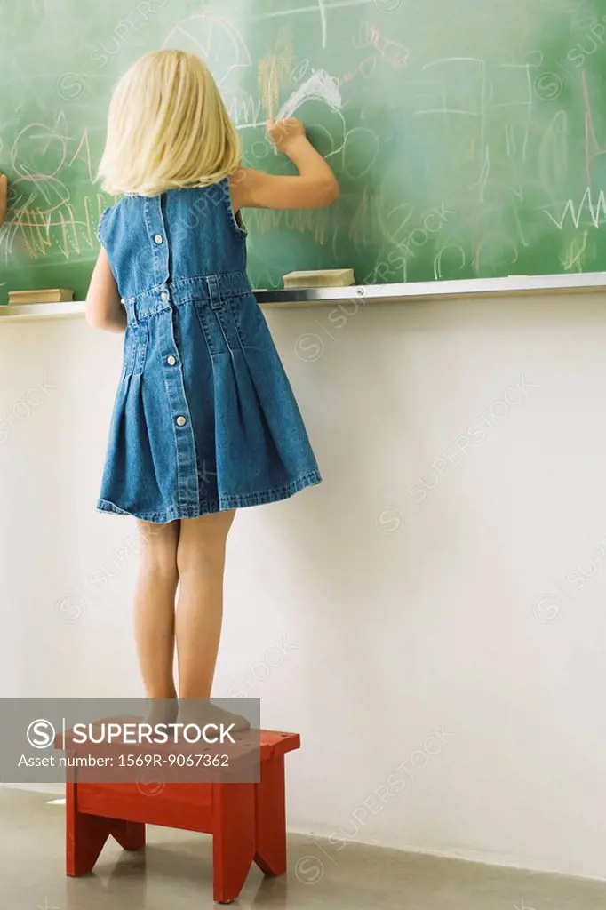 Little girl standing on stool, scribbling on blackboard, rear view