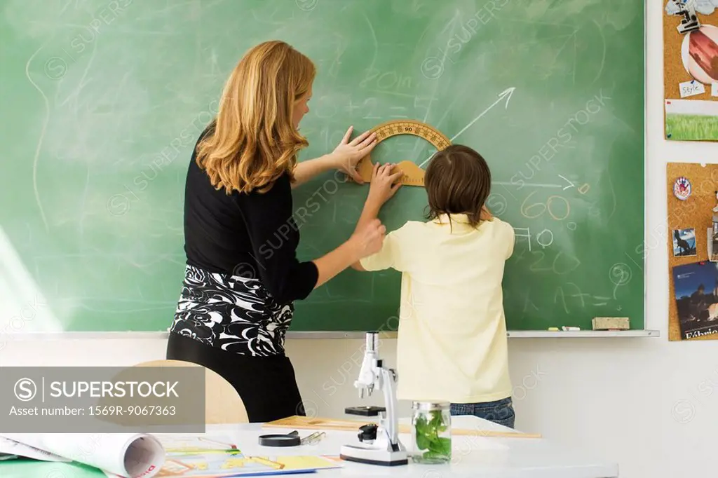 Teacher helping boy draw angle on blackboard using protractor, rear view