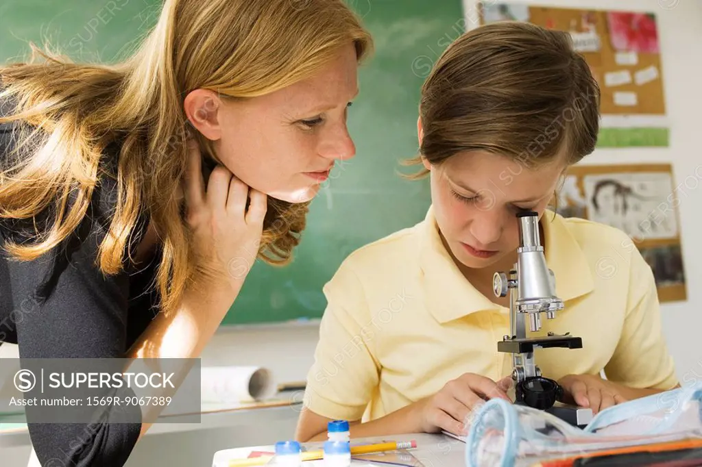 Boy looking through microscope, teacher watching
