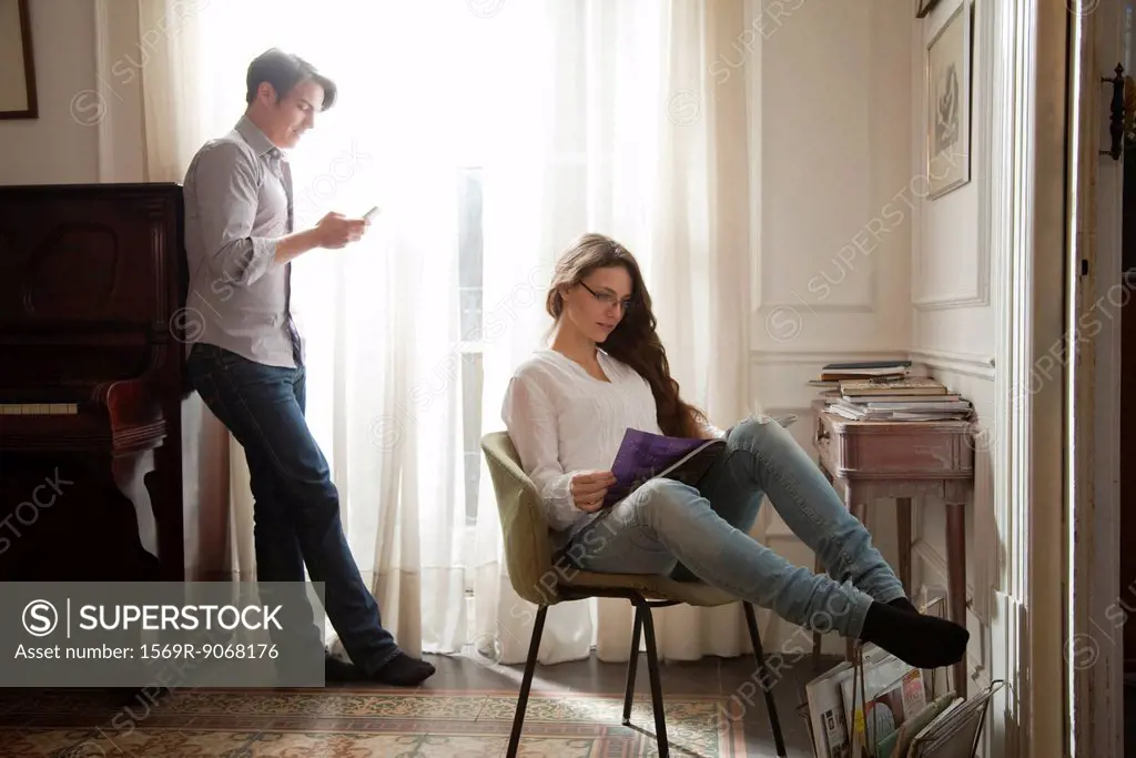 Couple relaxing at home, woman reading magazine, man looking at cell phone