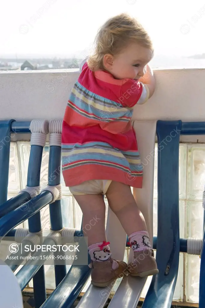 Toddler girl standing on chair, peeking over balcony railing, rear view