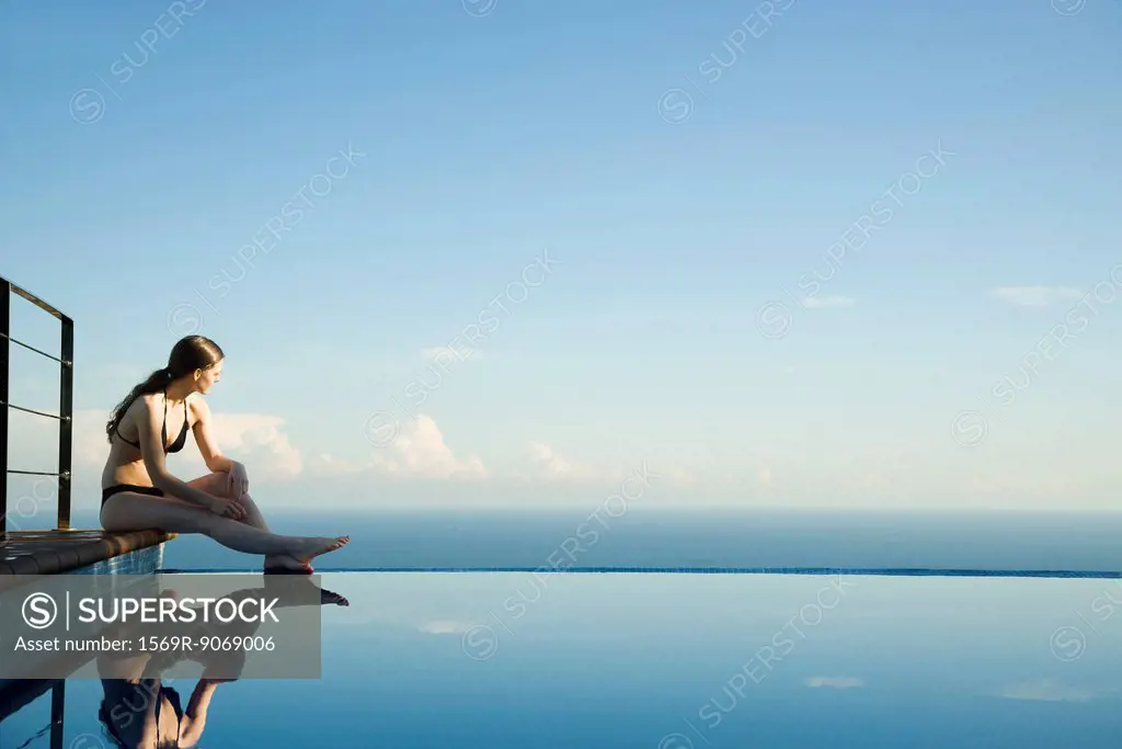 Woman sitting at edge of infinity pool, looking at view