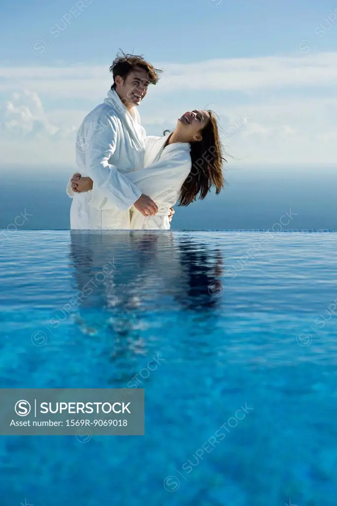 Couple embracing and laughing at edge of infinity pool, both wearing bathrobes