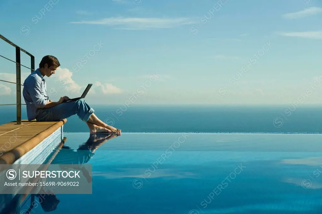 Man sitting beside infinity pool, using laptop computer