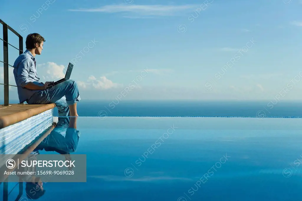 Man sitting at edge of infinity pool with laptop computer, looking at view