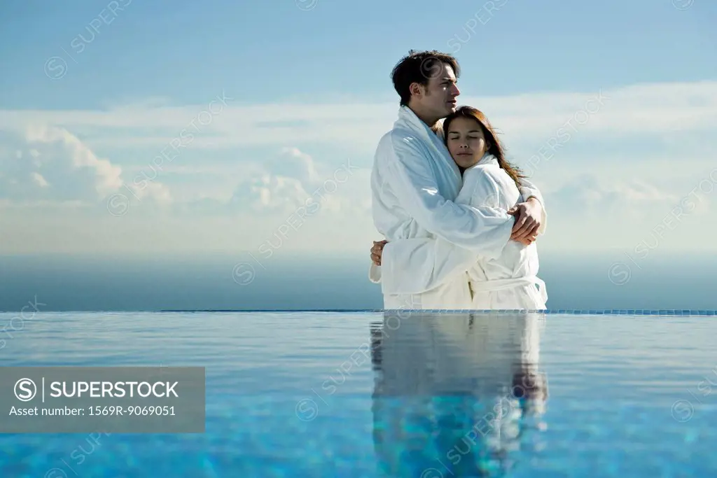 Couple embracing at edge of infinity pool, both wearing bathrobes