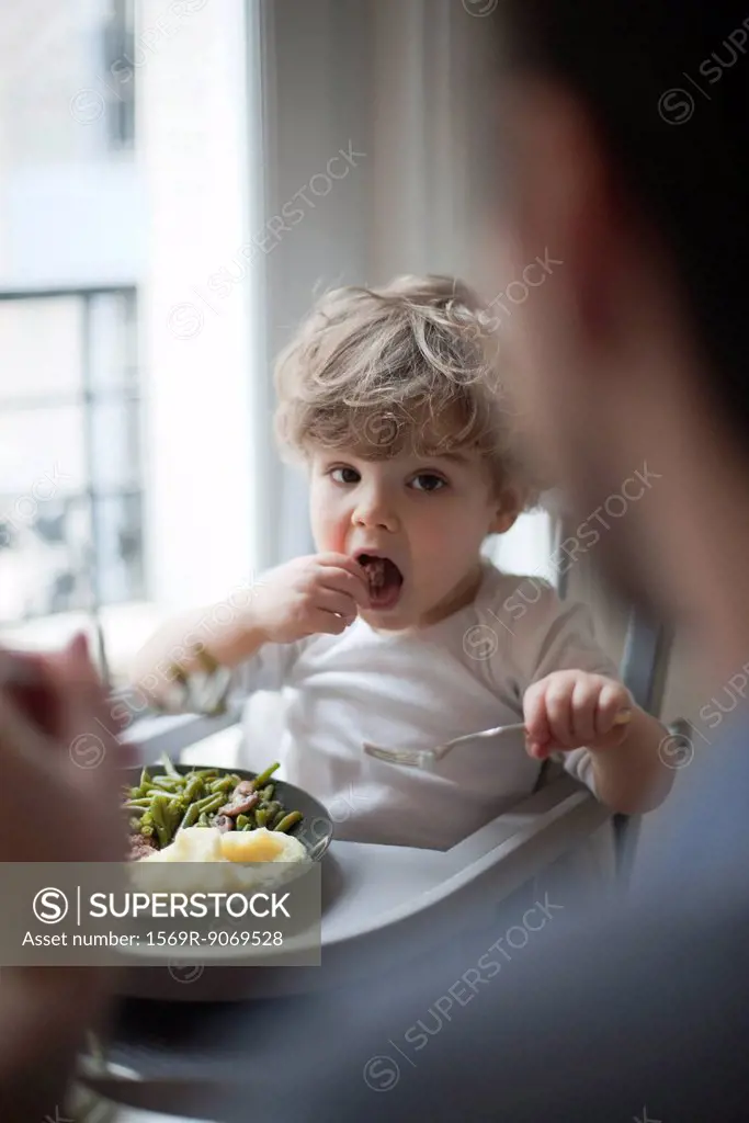 Toddler boy eating meal, looking at parent in foreground