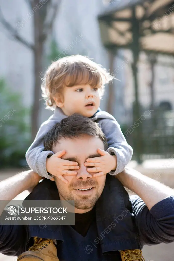 Father carrying toddler son on his shoulders, son covering father´s eyes with his hands