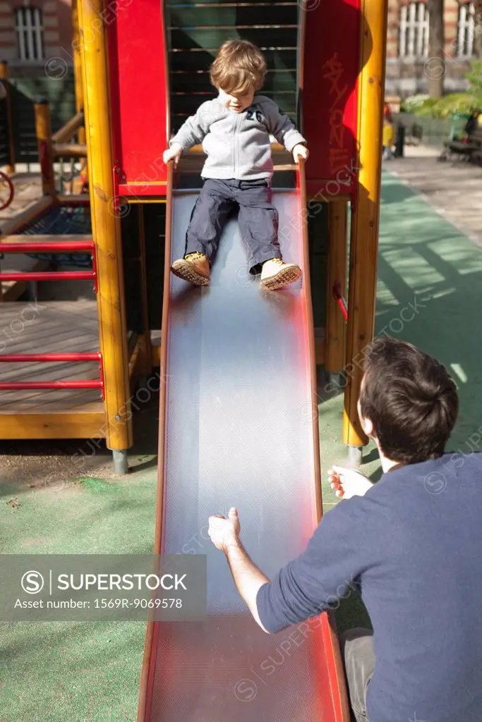 Toddler boy preparing to go down playground slide, father waiting at the bottom