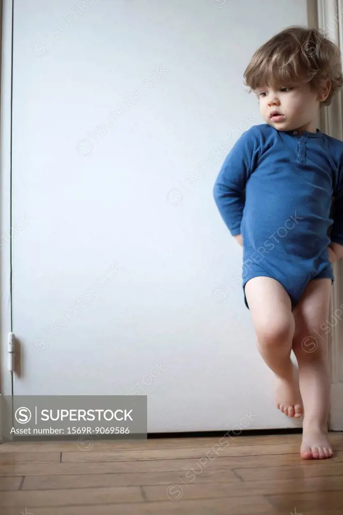 Toddler boy standing on one leg against door, looking away in thought, portrait
