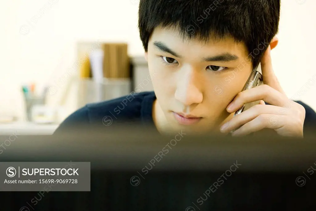 Young man looking at computer monitor and talking on cell phone
