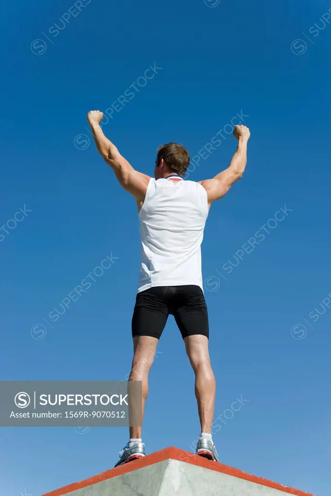 Male athlete standing on winner´s podium with arms raised in victory, rear view