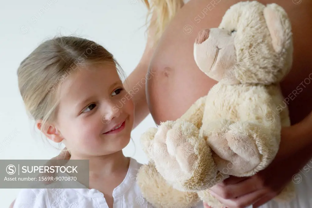 Girl beside pregnant mother, smiling at teddy bear