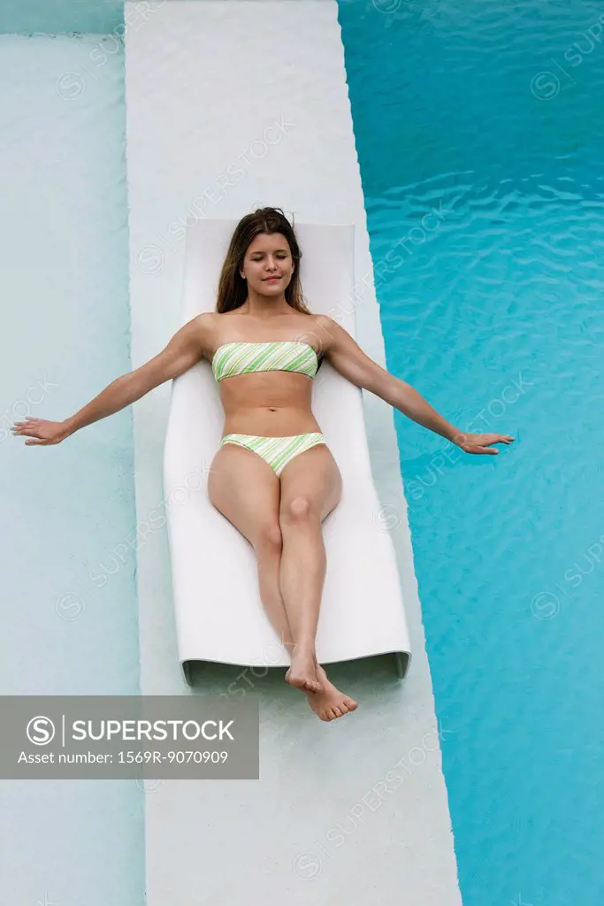 Young woman reclining on deck chair on walkway between swimming pools