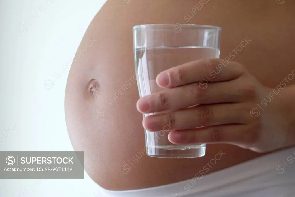 Woman holding glass of water beside pregnant belly, cropped