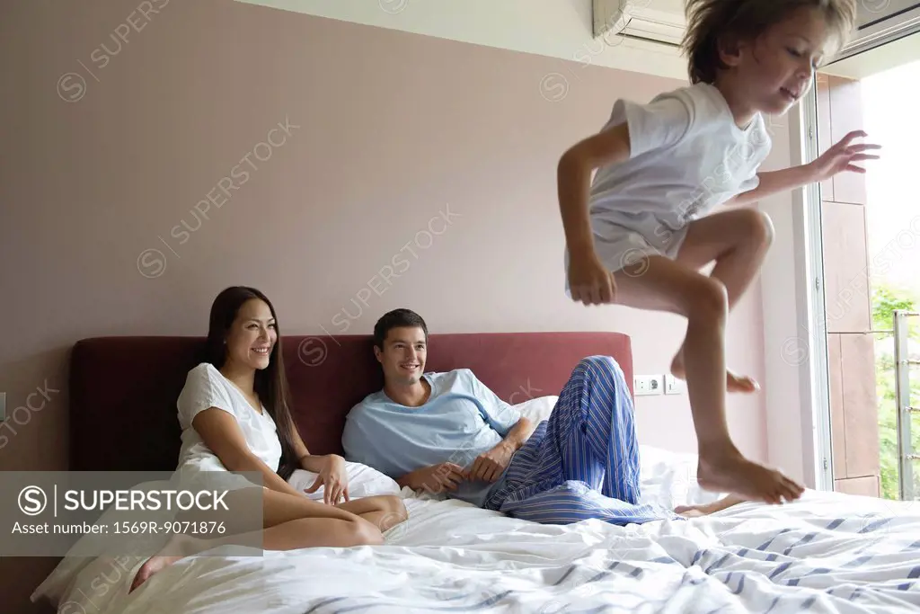 Parents watching as young son jumps on bed
